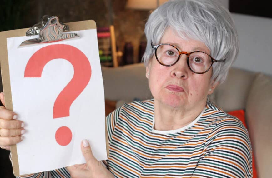 An older woman with gray hair and glasses holds up a clipboard displaying a large red question mark, sitting indoors on a couch.
